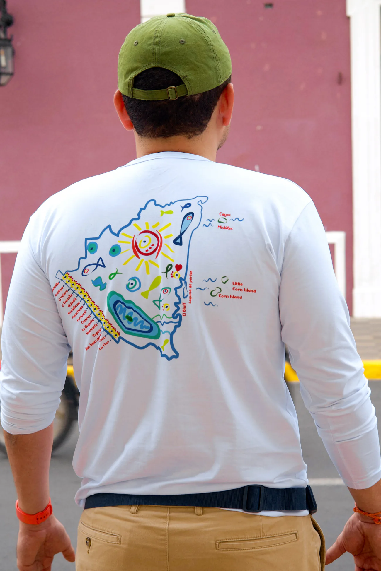 De espaldas en una calle de Granada, con gorra verde oliva y la camisa manga larga blanca con el mapa de Nicaragua dibujado a mano en azul, lleno de peces, un sol amarillo y un corazón rojo