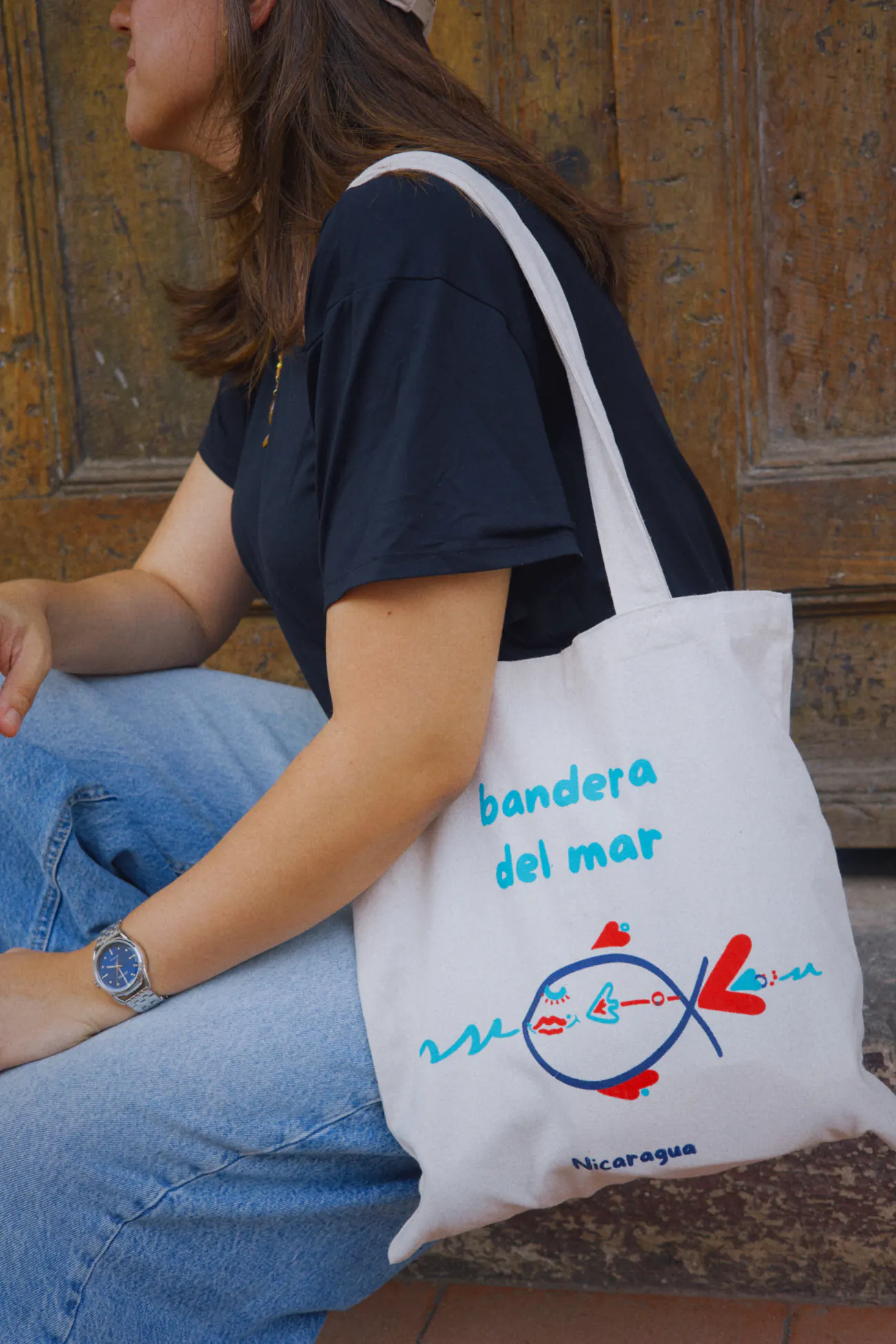 Primer plano de la totebag de lona con el pez azul y rojo y el nombre bandera del mar, colgada al hombro frente a un portón de madera en Granada