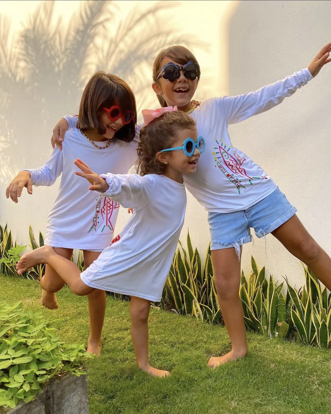 Tres nenas posando en el pasto con las camisetas manga larga blancas de la línea kids de bandera del mar y el pez pintado a mano en el frente