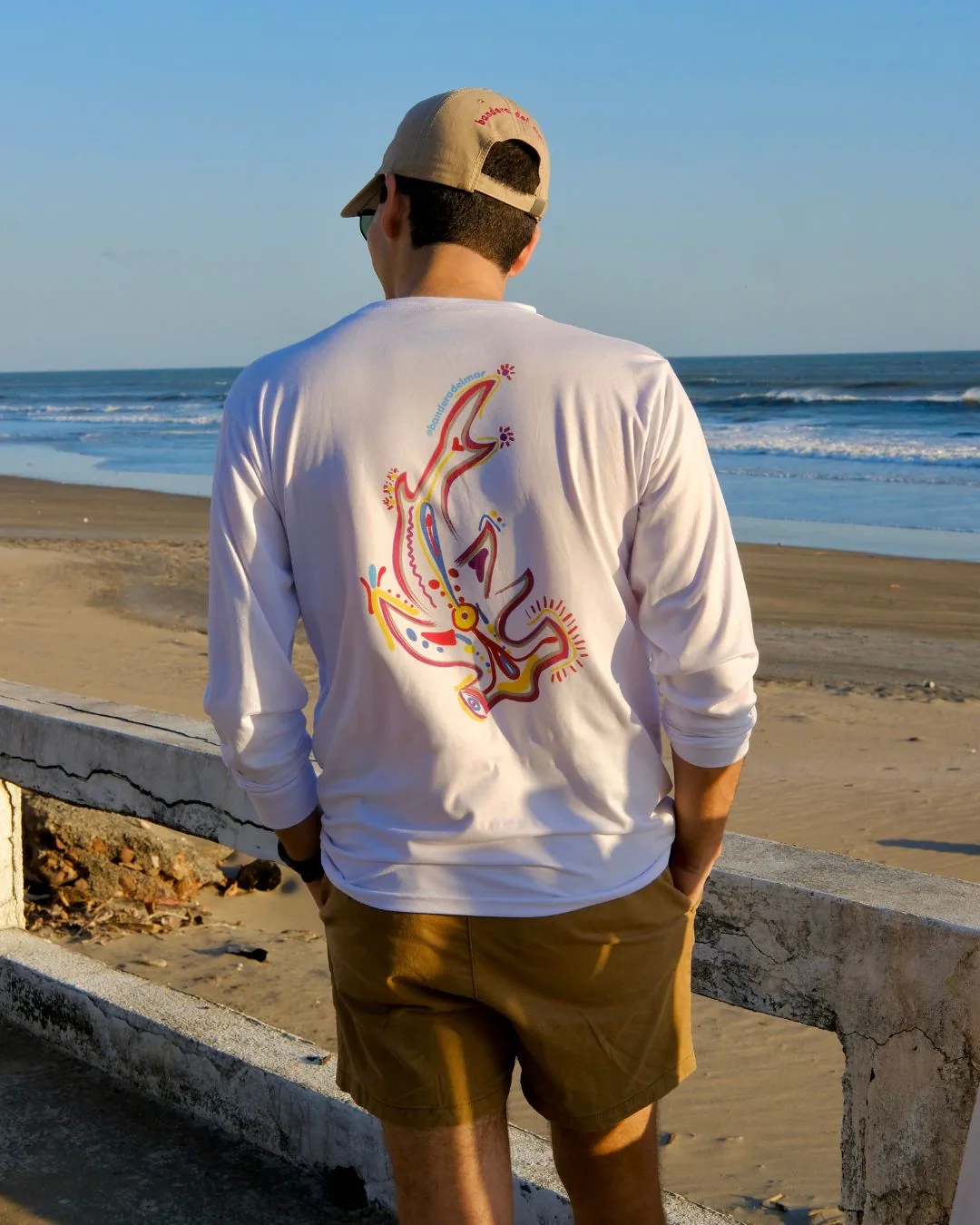 De espaldas frente al mar: el tiburón martillo pintado en la espalda de una camisa manga larga blanca, apoyado en el malecón