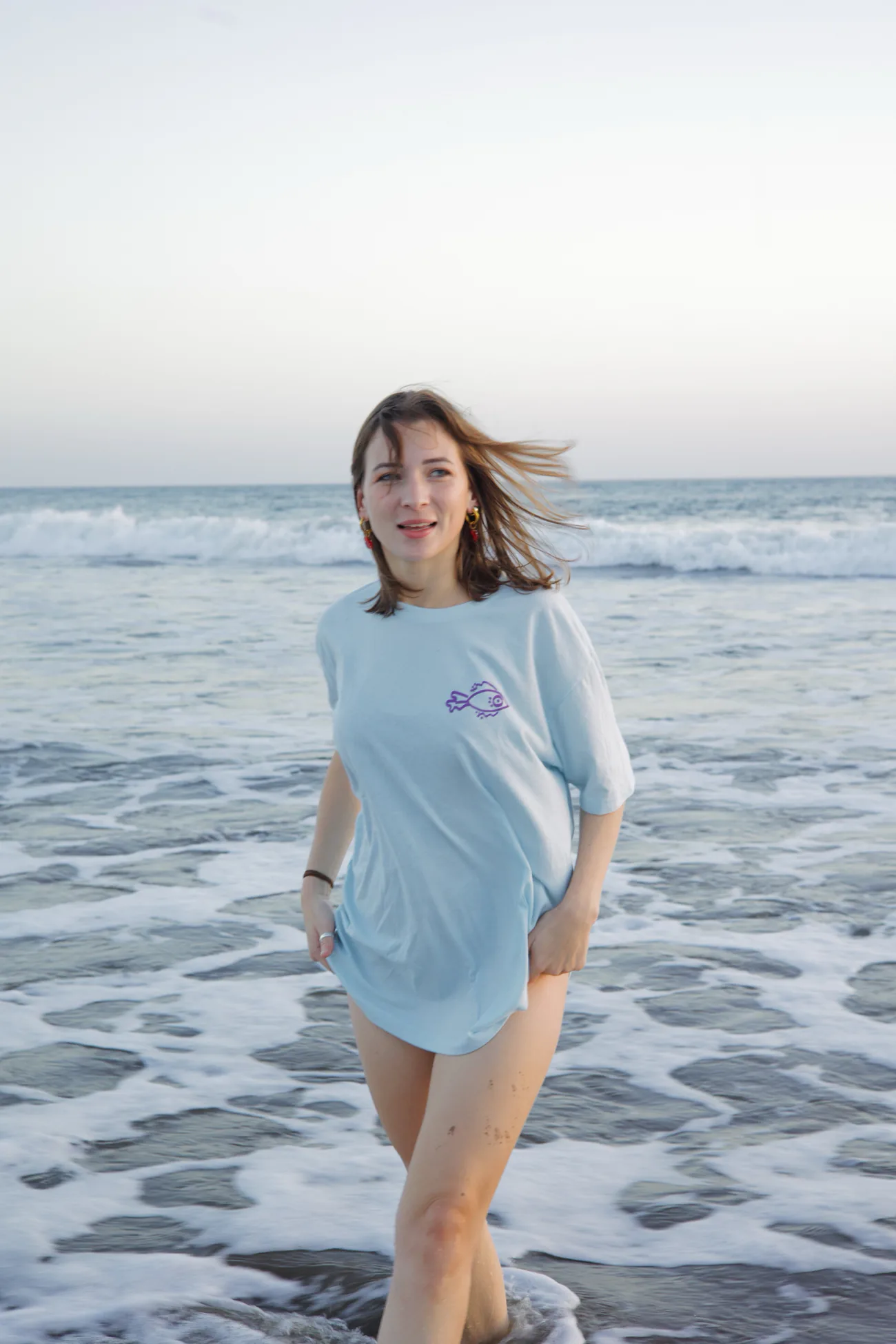 Parada en el agua, con la camiseta celeste y el pez morado pintado en el pecho, mientras rompe la ola atrás