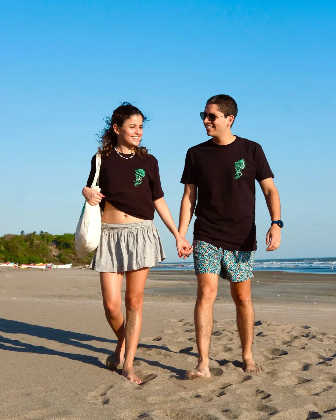 Una pareja camina descalza y de la mano por la arena, los dos con la medusa verde pintada en el pecho de la camiseta