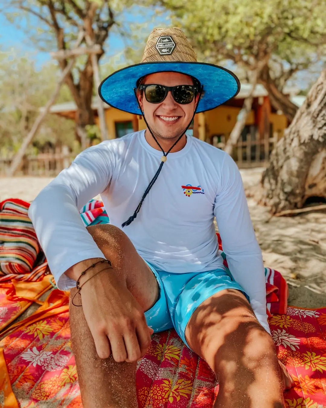 Sentado en la arena sobre una toalla tropical roja, con sombrero de palma de ala azul, la camisa manga larga blanca con el diseño de trazo a mano en el pecho y los shorts de baño turquesa