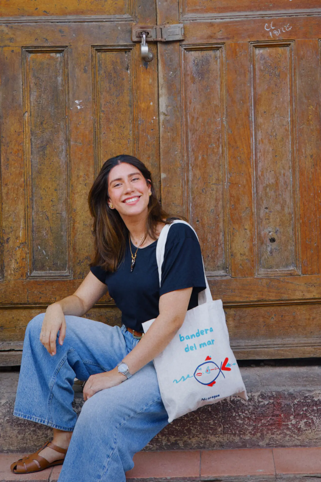 Sentada en el escalón de un portón de madera ocre, riéndose, con camiseta azul marino, jeans anchos y la totebag de lona del pez colgada al hombro